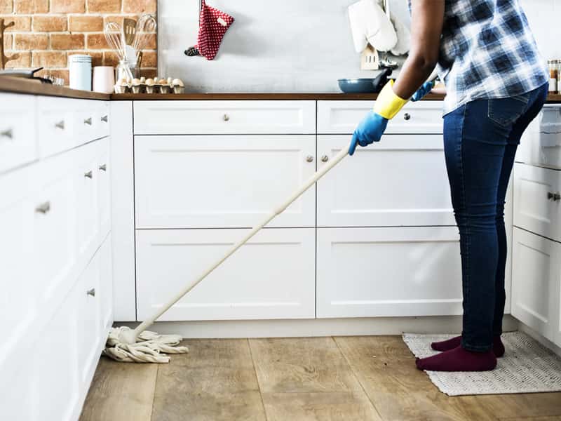 Une femme nettoie la cuisine, vêtue d’une chemise à carreaux, de gants bleus et d’un jean. Elle tient un balai pour passer la serpillière.