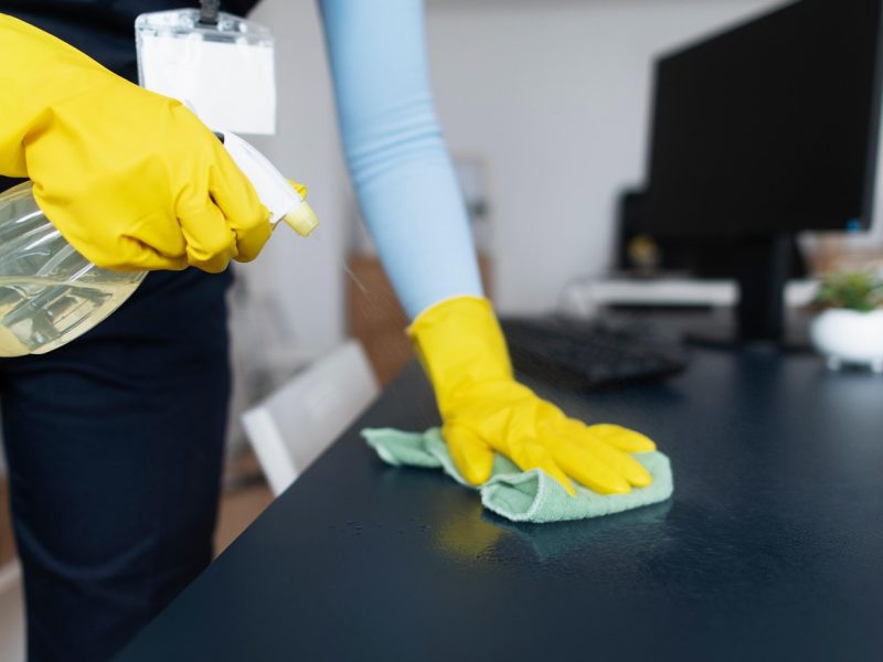 Une femme en train de nettoyer un bureau, portant des gants jaunes et tenant un produit de nettoyage dans sa main.